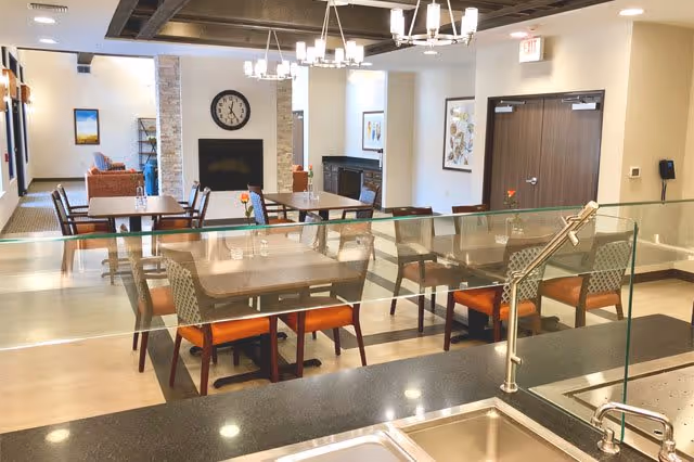 Interior view of a dining area in a senior living facility with multiple tables and chairs, a fireplace with a clock above it, and modern lighting fixtures. The photo is taken from behind a kitchen counter with a sink and glass partition.