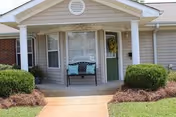 Front porch entry of a single-story senior living residence with white columns, a bench, a green door, and trimmed shrubs.