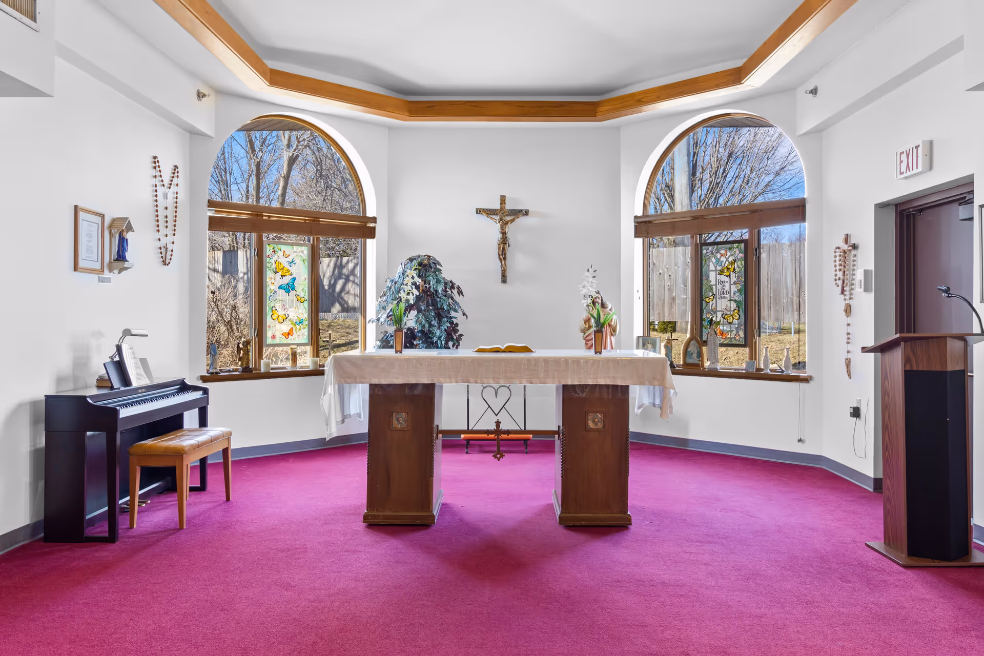 Interior of a small chapel or prayer room with a wooden altar covered by a white cloth in the center, a crucifix mounted on the wall behind it, two large arched windows with stained glass butterfly designs, a piano with a bench on the left, and a wooden lectern on the right. The room has a bright magenta carpet and white walls with wooden trim on the ceiling.