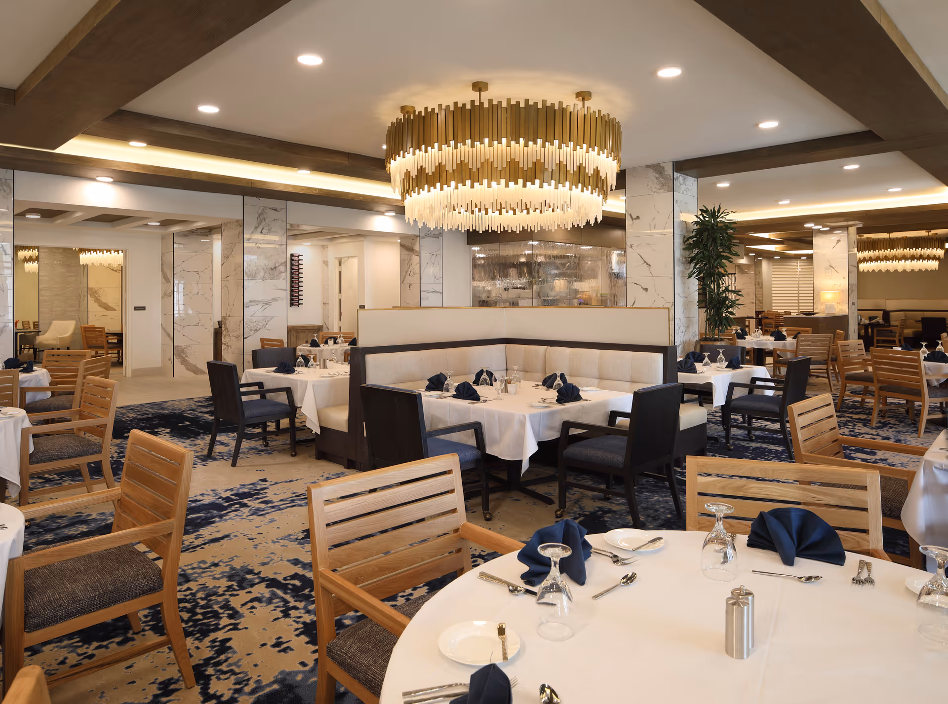 A spacious and elegant dining room with multiple tables set with white tablecloths, navy blue napkins, glassware, and silverware. The room features wooden chairs, a large modern chandelier, marble columns, and a patterned carpet with blue and beige tones.