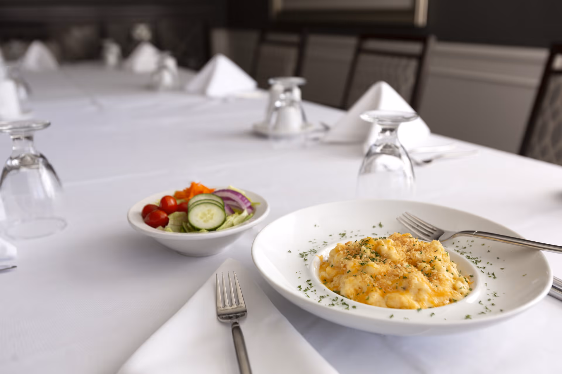 A white tablecloth-covered dining table set with upside-down glasses and folded white napkins. In the foreground, there is a plate of macaroni and cheese garnished with herbs and a small bowl of salad containing cherry tomatoes, cucumber slices, red onion, and shredded carrots. Silverware is placed on white napkins beside the plates.