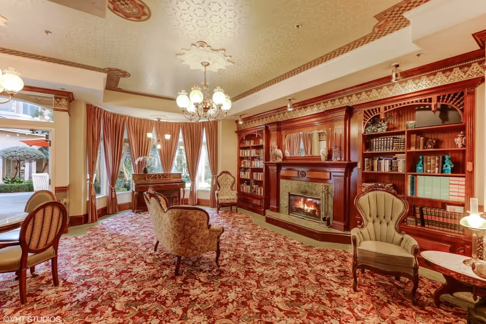 A richly furnished sitting room with a fireplace flanked by built-in bookshelves, upholstered chairs and sofas, a piano, and an ornate chandelier.