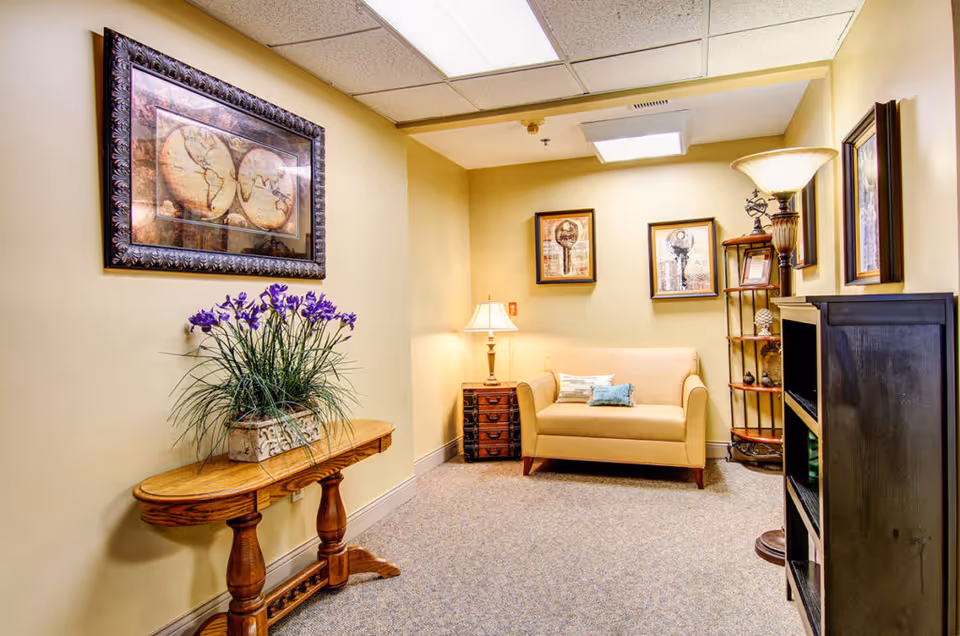 Small furnished sitting area with a beige loveseat, side table and lamp, framed artwork, a wooden console table with flowers, and shelving.