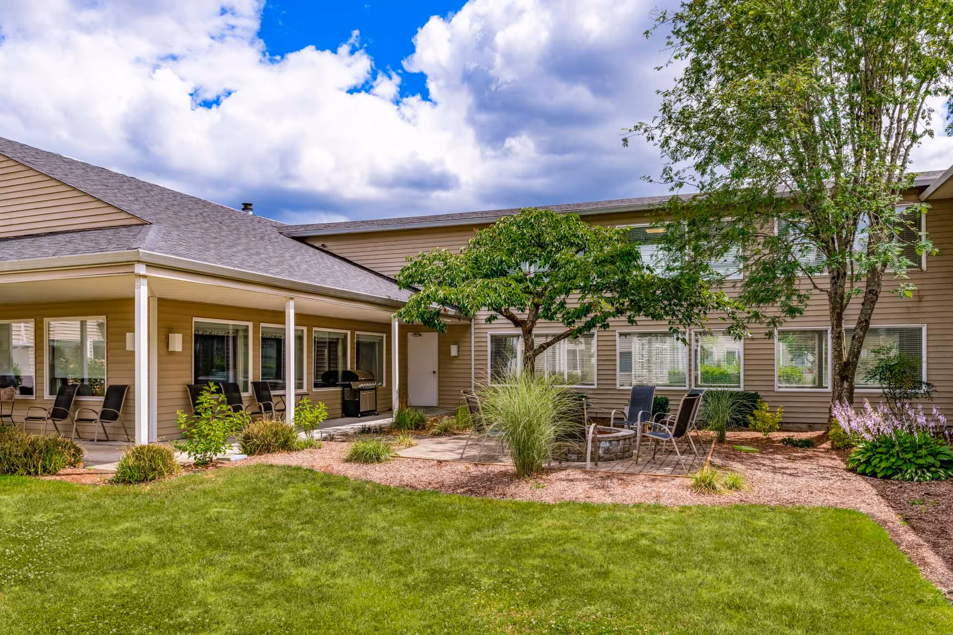 Outdoor courtyard area of Holiday Edgewood Downs featuring a green lawn, trees, shrubs, patio chairs arranged around a fire pit, and a covered porch with additional seating and a grill under a partly cloudy sky.