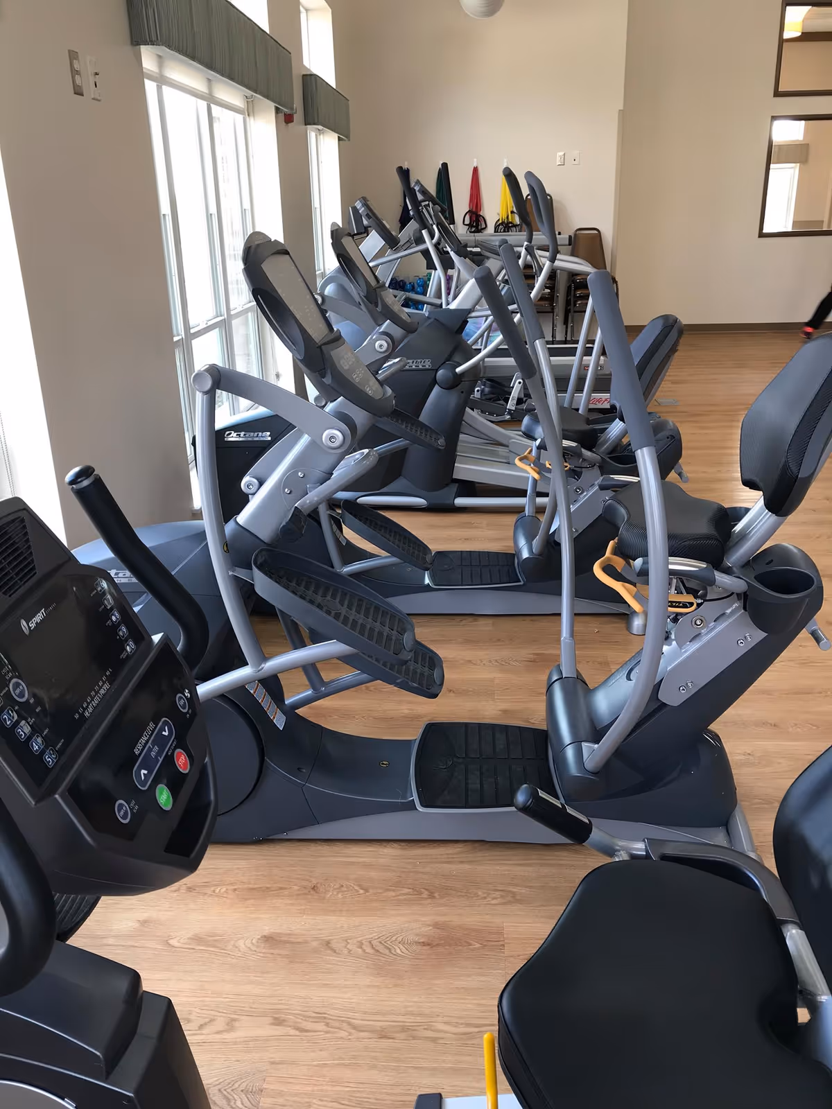 Interior view of a fitness room with several elliptical machines and exercise equipment lined up next to large windows with natural light. The room has wooden flooring and light-colored walls.