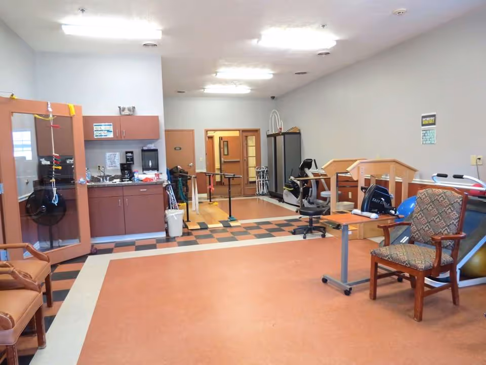 A rehabilitation room with exercise equipment including a stationary bike, parallel bars, and therapy chairs. The room has a checkered floor section near a kitchenette area with cabinets, a sink, and a coffee maker. There are chairs and a small table in the foreground, and the walls are painted light gray.