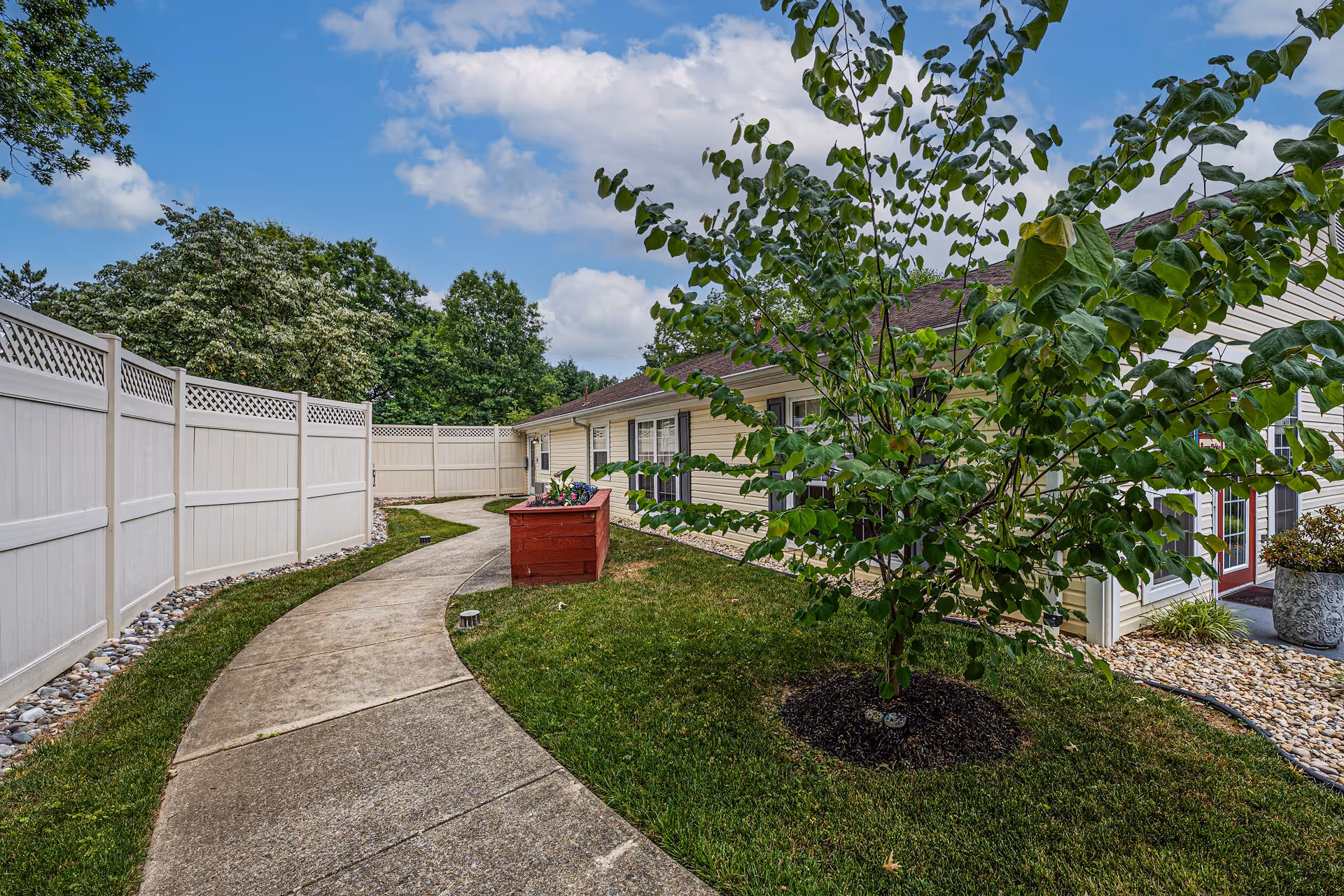 A curved concrete walkway bordered by grass and a white privacy fence leads to a single-story building with beige siding and dark shutters. There is a small tree and a red planter box with flowers along the path. The sky is blue with scattered clouds.