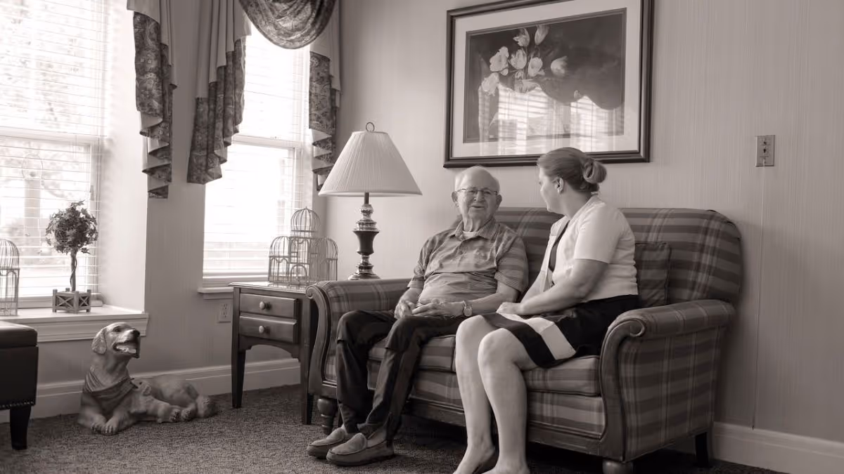 An elderly man and a woman sitting and talking on a plaid sofa in a well-lit living room with large windows, a side table with a lamp, decorative birdcages, a framed floral picture on the wall, and a dog statue on the floor near the window.