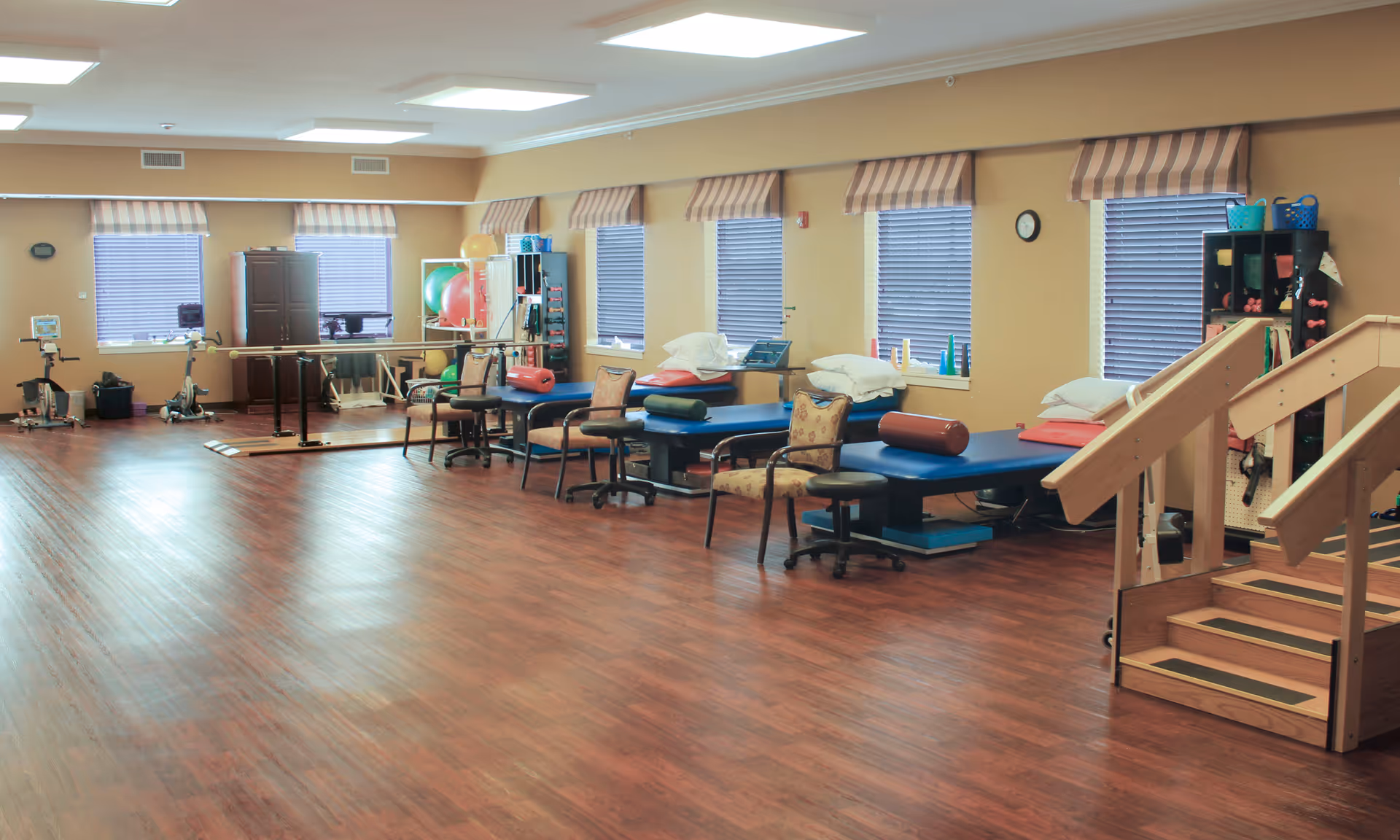A spacious physical therapy or rehabilitation room with wooden flooring, multiple therapy tables with pillows and bolsters, chairs, exercise equipment including stationary bikes, therapy balls, and parallel bars. There are windows with striped valances and a set of wooden stairs with handrails for mobility training.