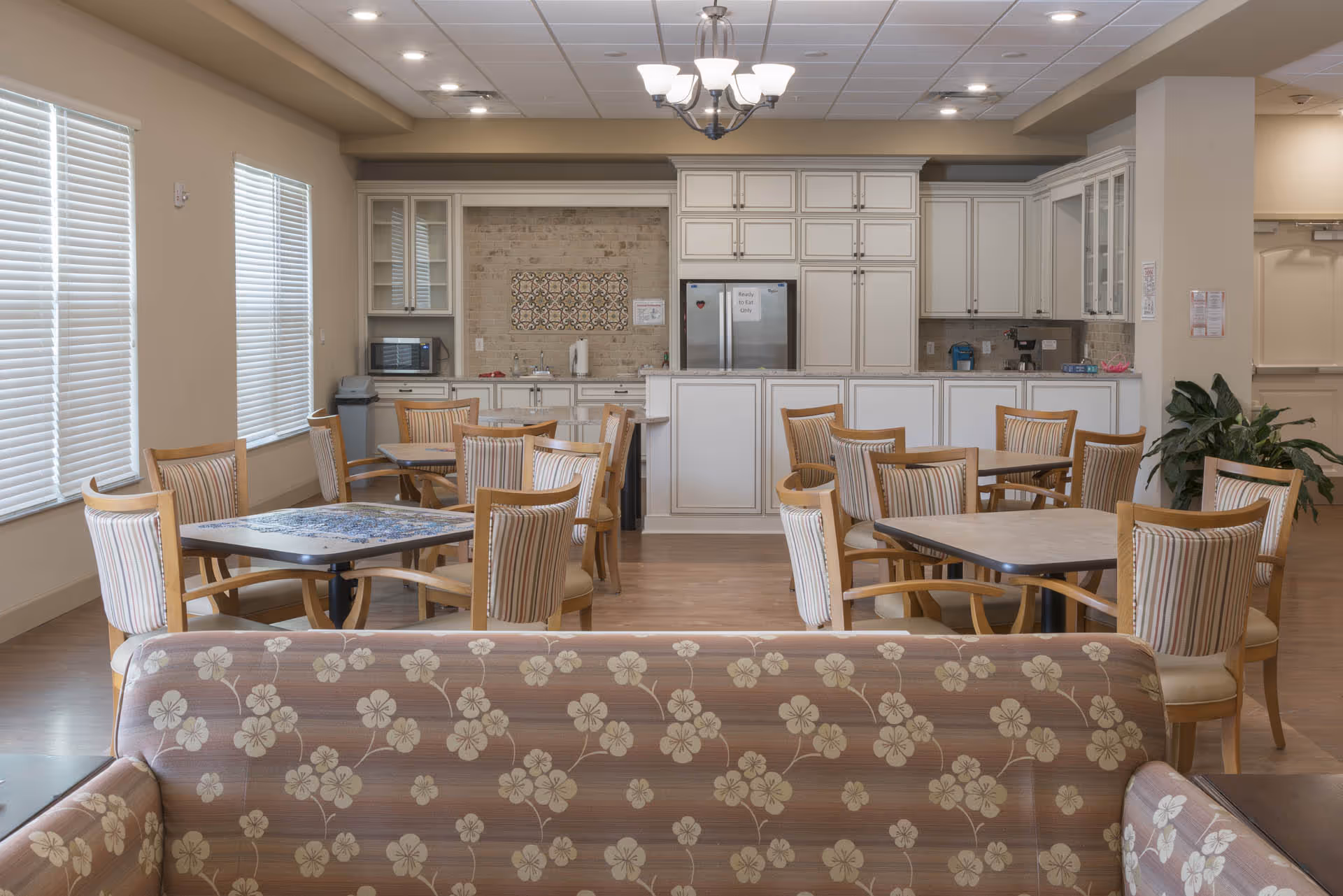 A bright and clean dining area in a senior living facility with multiple tables and chairs arranged neatly. The room features large windows with blinds, a floral patterned couch in the foreground, and a kitchen area with white cabinetry, a refrigerator, microwave, and coffee maker in the background. A chandelier hangs from the ceiling.