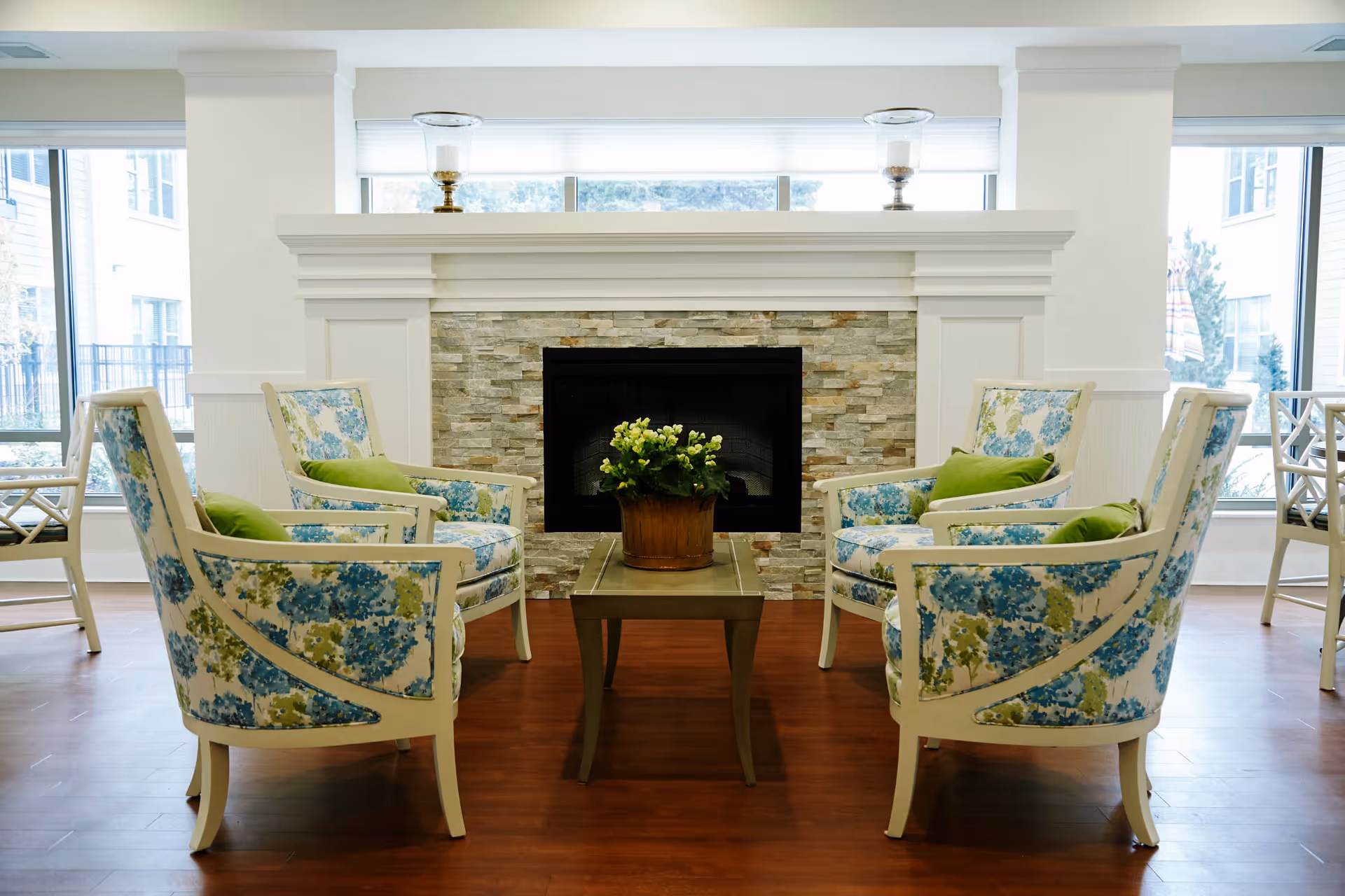 A cozy seating area with four floral-patterned armchairs arranged around a small wooden coffee table with a potted plant. Behind the seating area is a fireplace with a stone surround and white mantel, flanked by large windows letting in natural light.