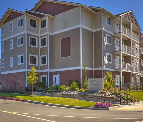 Exterior view of a multi-story senior living facility building with beige and brown siding, multiple windows, balconies, and landscaped greenery including small trees and flowers. A sign in front reads 'Vintage at Lakewood'.