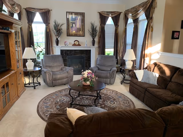 Living room with two gray recliners and brown sofas centered around a round coffee table and a fireplace beneath curtained windows.