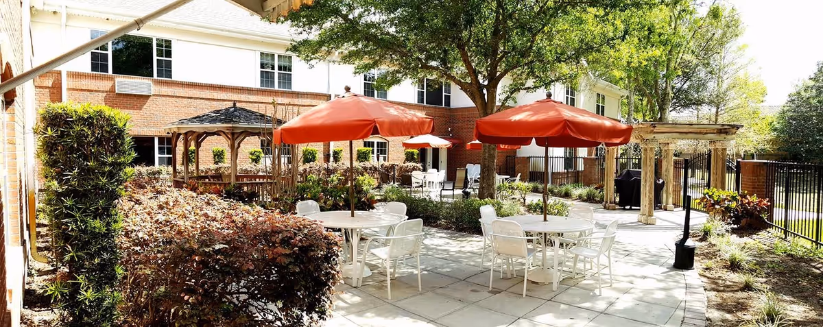 Outdoor patio area at Ambiance at Maitland featuring round white tables with red umbrellas, white chairs, a wooden gazebo, and a pergola surrounded by greenery and trees, adjacent to a brick building.