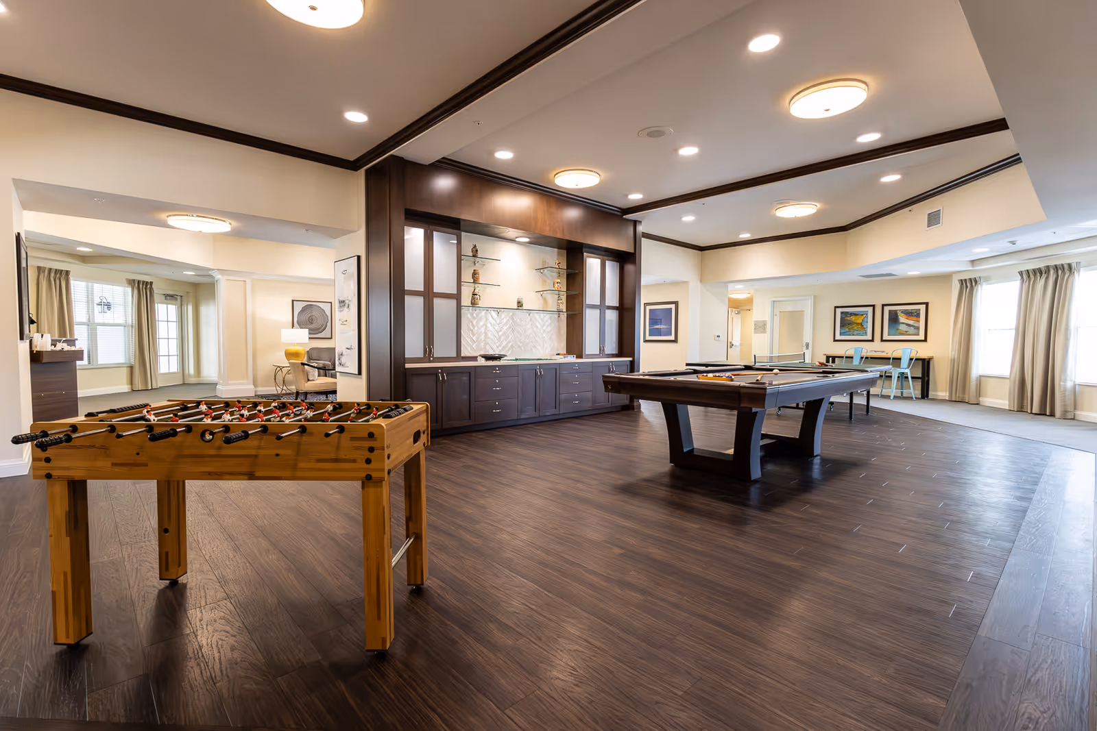 A spacious game room with a foosball table in the foreground and a pool table in the center. The room features dark wood flooring, built-in cabinetry with decorative items, and several framed artworks on the walls. Large windows with beige curtains allow natural light to fill the space. There is also a small seating area with a table and chairs near the windows.