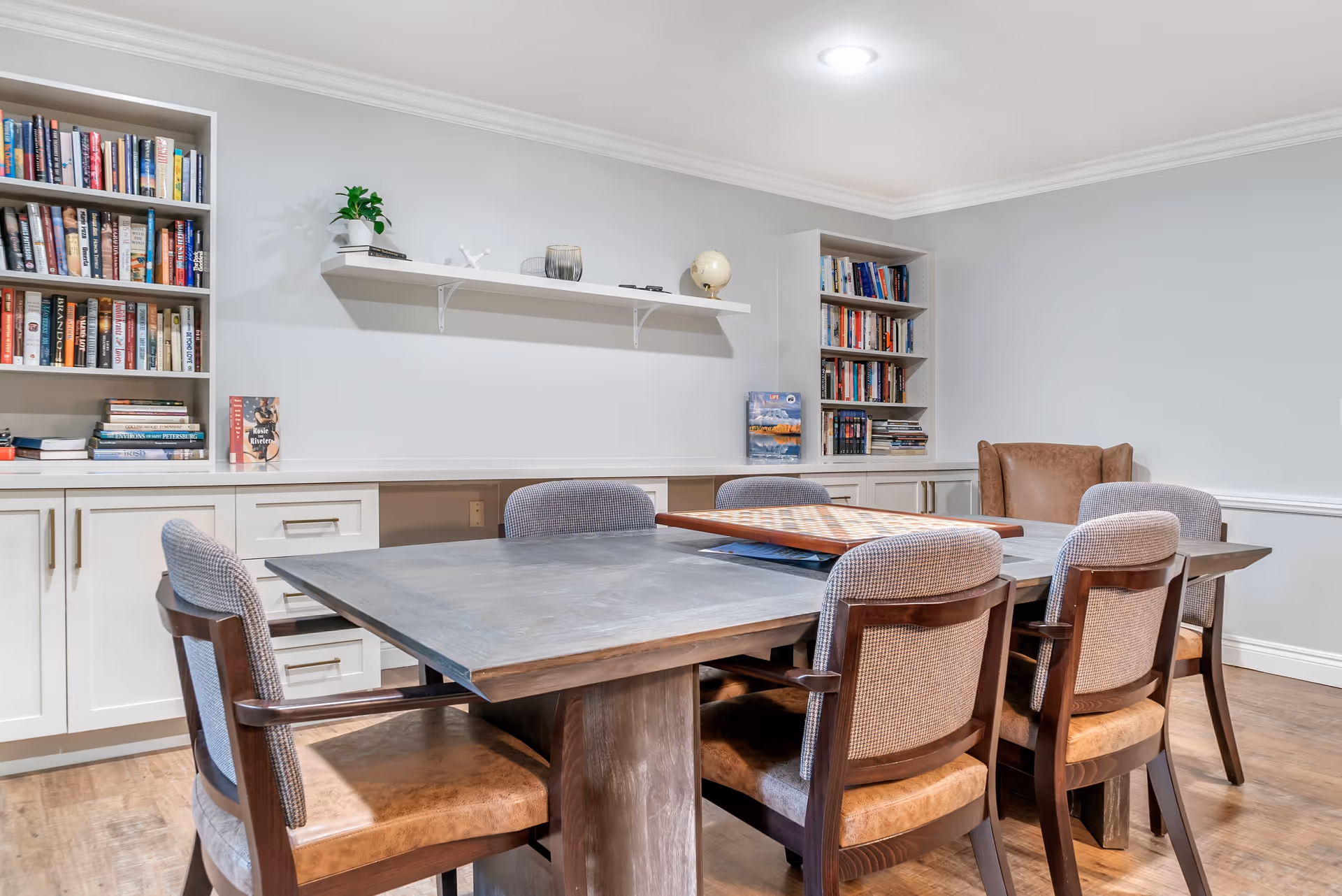 A cozy room with a large wooden table surrounded by six cushioned chairs. The table has a chessboard on it. The room features built-in white cabinets and bookshelves filled with books. A floating shelf on the wall holds decorative items including a small globe and a plant. The walls are painted light gray and the floor is wooden.