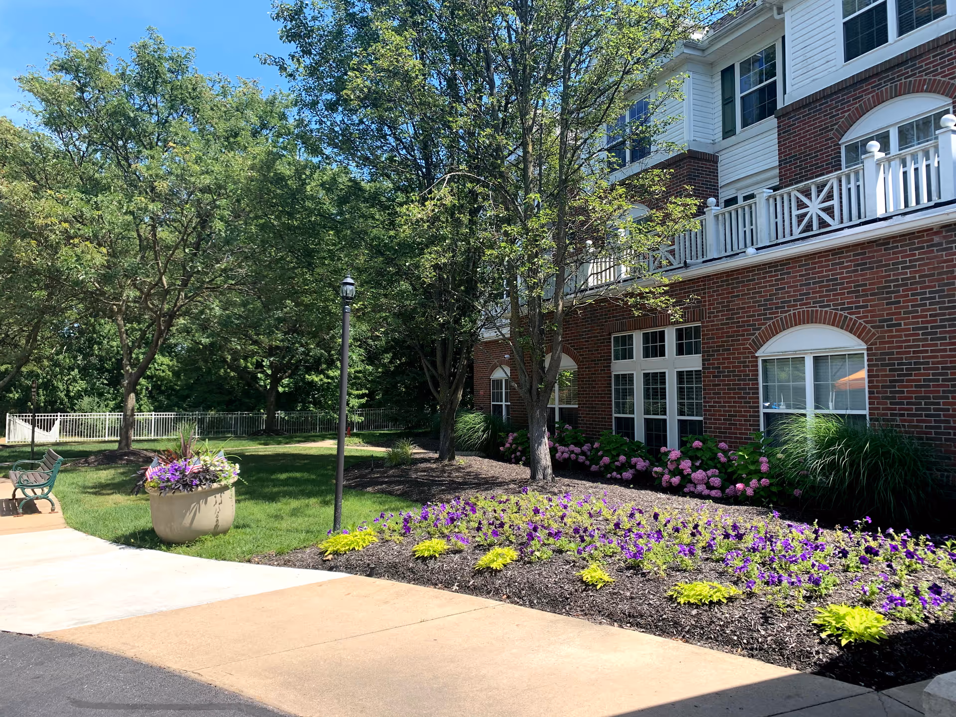 Exterior view of a senior living facility with a brick facade, white trim, and multiple windows. The foreground features a landscaped garden with purple and yellow flowers, green shrubs, and trees. There is a paved walkway, a bench, a lamppost, and a hammock in the grassy area under a clear blue sky.
