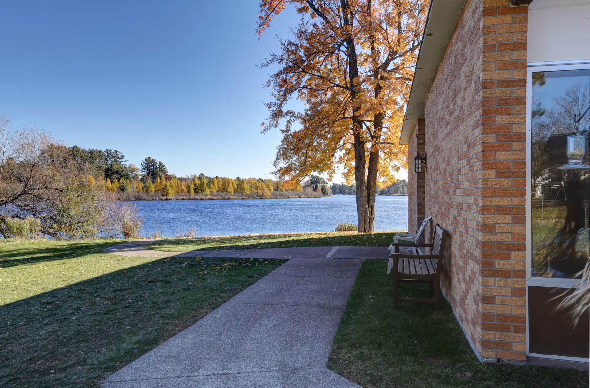 Brick-sided building exterior with benches and a paved path leading to a lake lined with autumn trees.