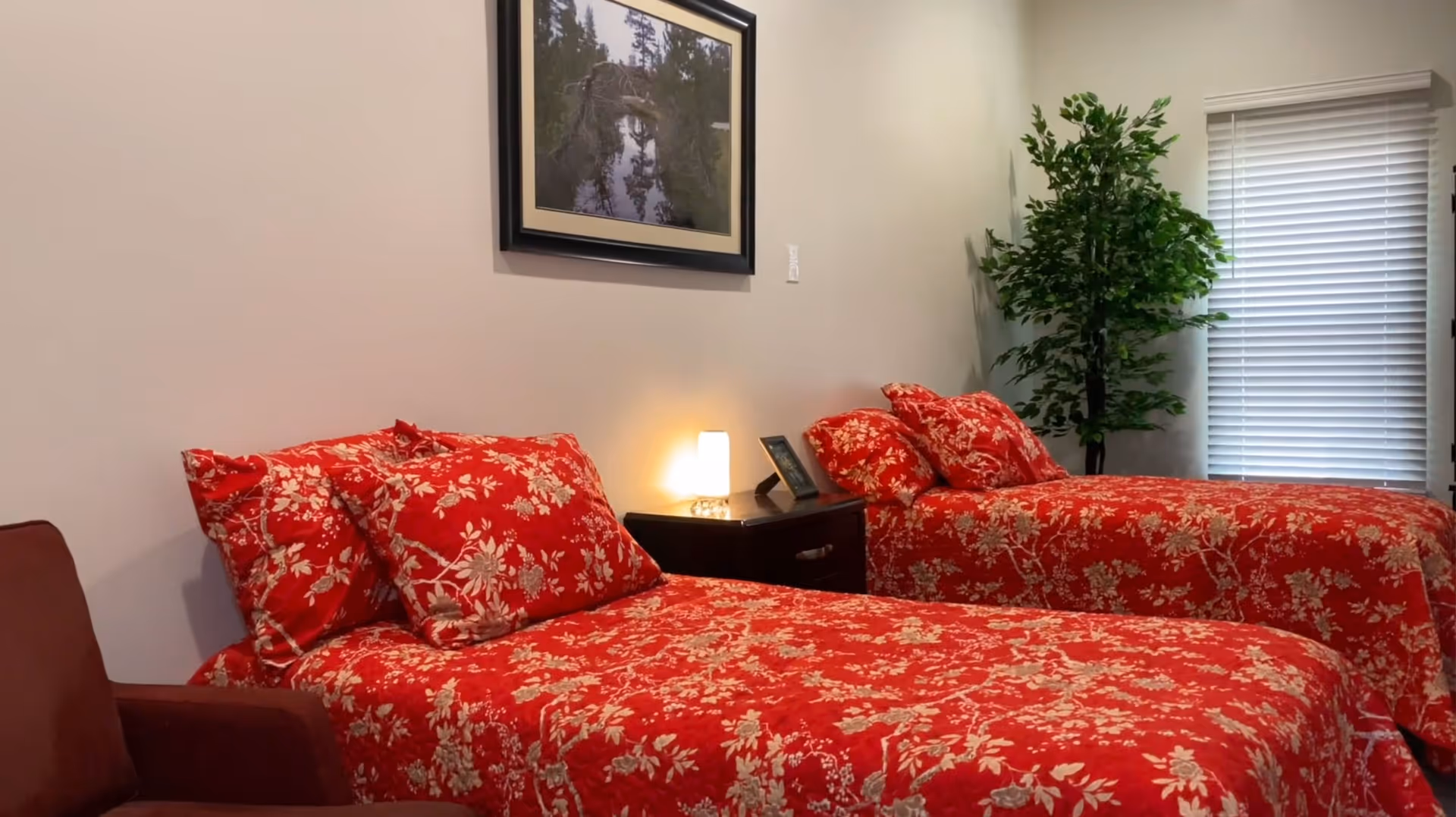 Two twin beds with red floral bedding in a bedroom with a nightstand, lamp, framed picture, and potted plant.