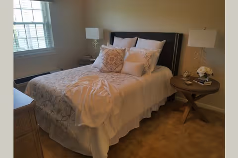 A cozy bedroom with a neatly made bed featuring multiple decorative pillows and a light-colored blanket. There are two bedside tables, each with a lamp, and one table also has a small clock and a vase with flowers. A window with blinds allows natural light into the room.
