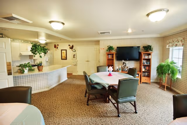 A cozy assisted living common area featuring a dining table with six chairs, a flat-screen TV mounted on the wall, two wooden bookshelves, and several potted plants. The room has patterned wallpaper, carpeted flooring, and a window with floral curtains allowing natural light to enter. A kitchen area with white cabinets and a counter is visible in the background.