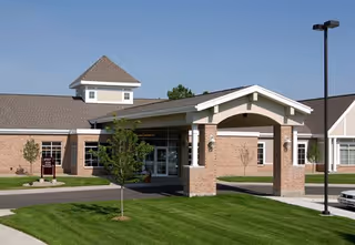 Exterior view of a brick senior living facility entrance with a covered porte-cochere and green lawn.