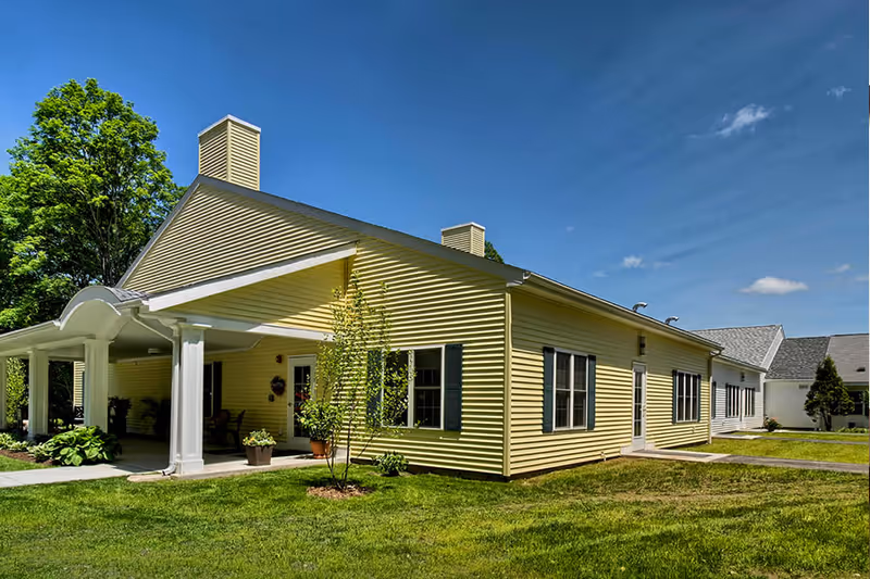 Exterior view of a single-story yellow building with white trim and a covered entrance supported by white columns, surrounded by green grass and trees under a clear blue sky.