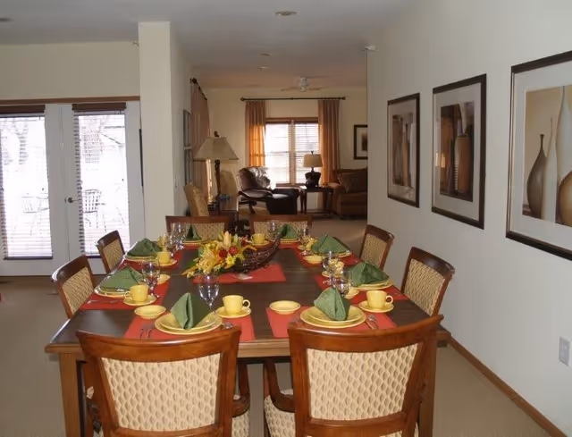 A dining room set with a rectangular wooden table and eight chairs with patterned upholstery. The table is set with yellow plates, cups, green folded napkins, wine glasses, and red placemats. A floral centerpiece is in the middle of the table. In the background, there is a living room area with a sofa, armchair, lamps, and a window with curtains. Three framed pictures hang on the wall to the right. Double glass doors with blinds are visible on the left side.