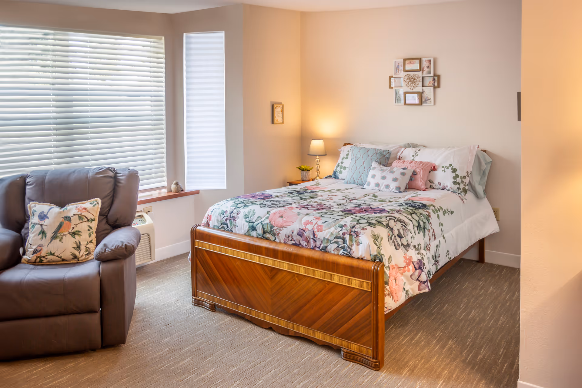 A bright, neatly decorated bedroom with a floral-patterned bed, wooden bedframe, recliner chair, and bedside lamp.