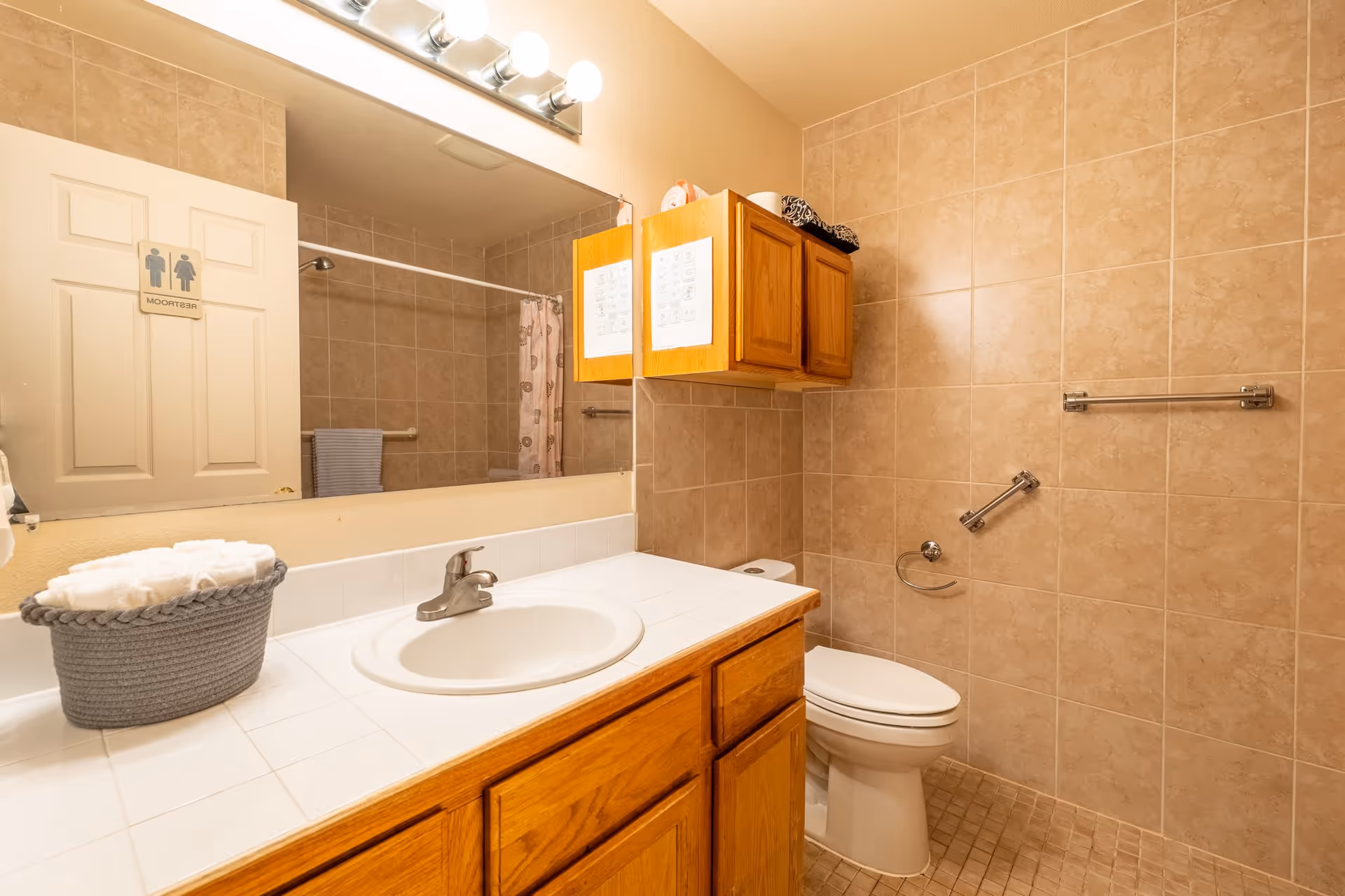 A clean bathroom with beige tiled walls and floor, featuring a white sink with a wooden cabinet below, a toilet, a large mirror above the sink, a wooden wall cabinet, a towel rack, and a shower area with a curtain. A basket with rolled white towels is placed on the counter. A restroom sign is visible on the door.