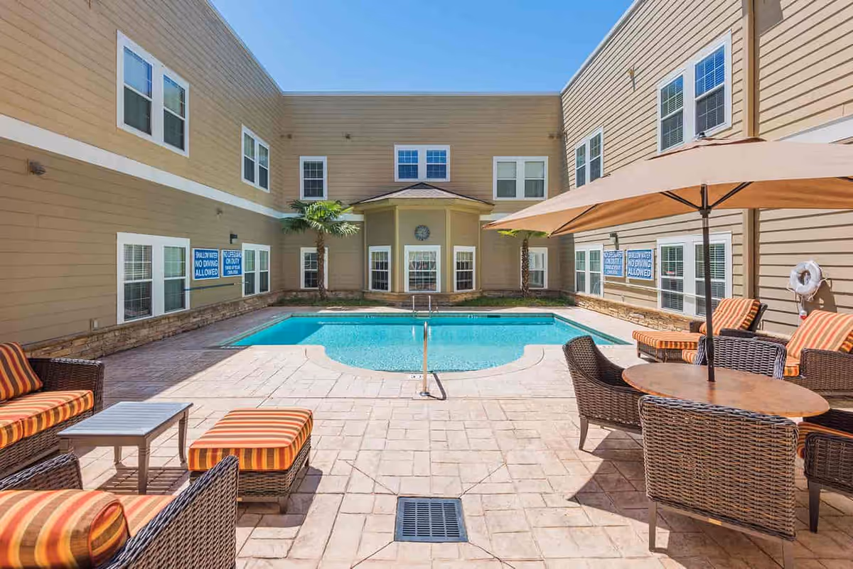 Sunlit courtyard pool area with wicker patio furniture, umbrellas, and a beige multi-story building surrounding the pool.
