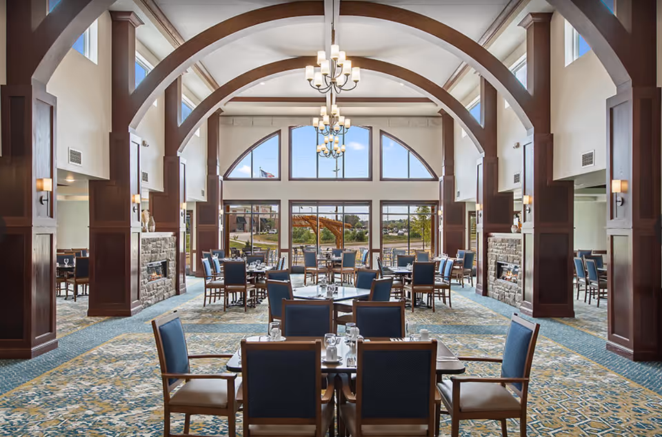 A spacious dining room in The Meadows Senior Living with multiple tables and chairs arranged neatly. The room features high ceilings with large arched windows allowing natural light to fill the space. There are elegant chandeliers hanging from the ceiling and stone fireplaces on either side of the room. The carpet has a patterned design, and the overall ambiance is warm and inviting.