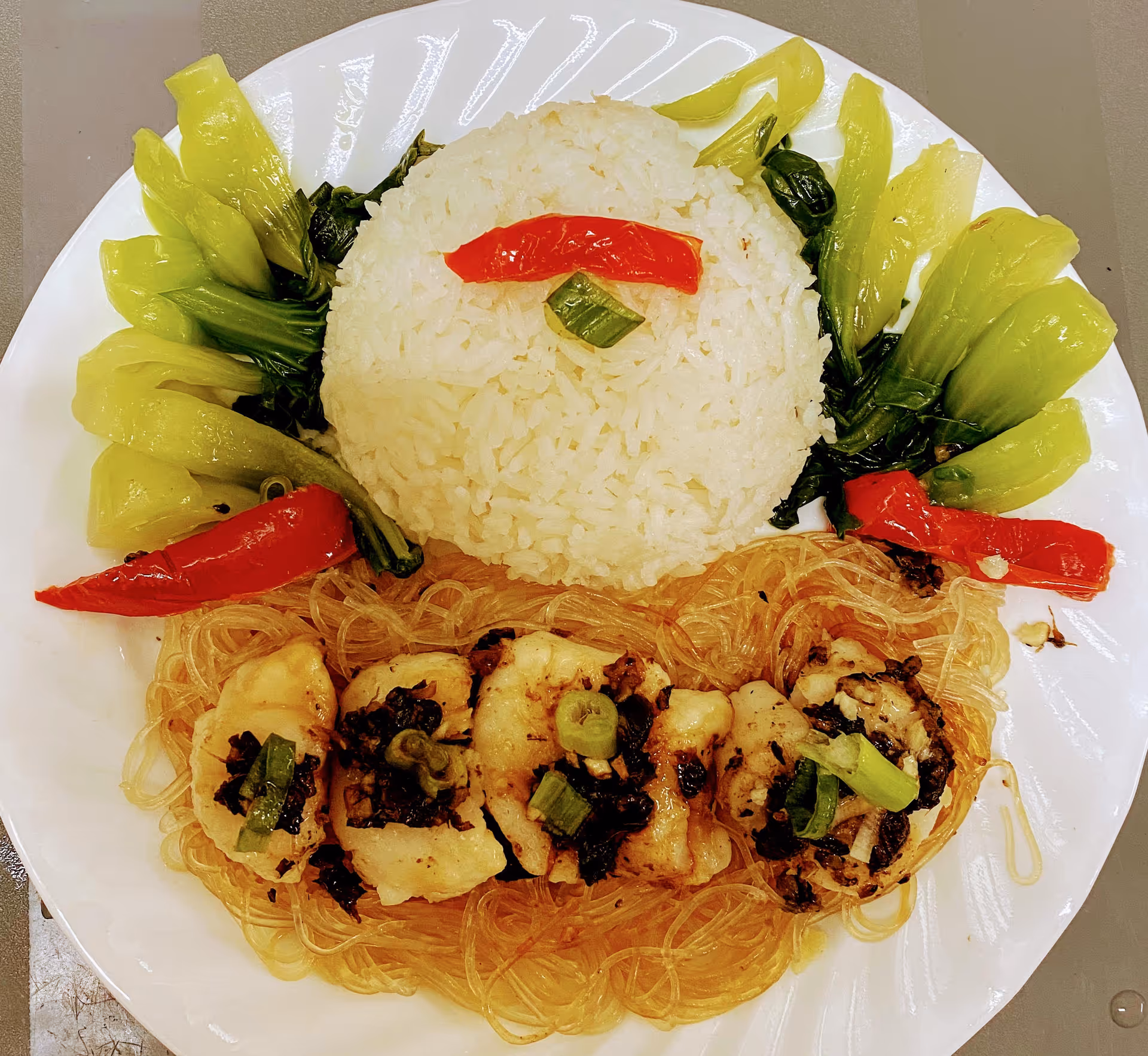 A plate of food featuring a mound of white rice garnished with a slice of red pepper and green onion on top, surrounded by cooked green leafy vegetables and red pepper slices. Below the rice, there are pieces of cooked fish or chicken with chopped green onions on a bed of clear noodles.