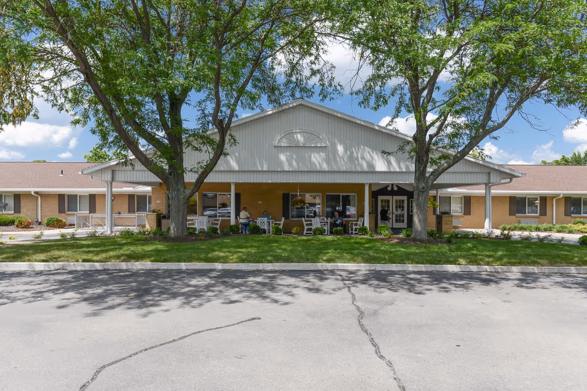 Front view of a single-story brick senior living facility with a covered entrance, porch seating, and large trees.
