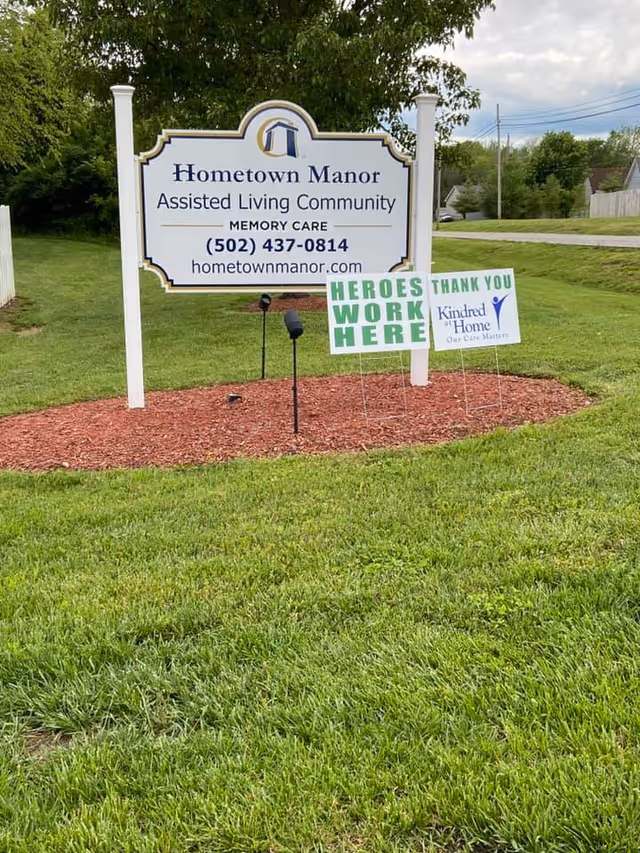 Outdoor sign for Hometown Manor Assisted Living Community on a grassy lawn with smaller 'Heroes Work Here' and 'Thank You' signs nearby.