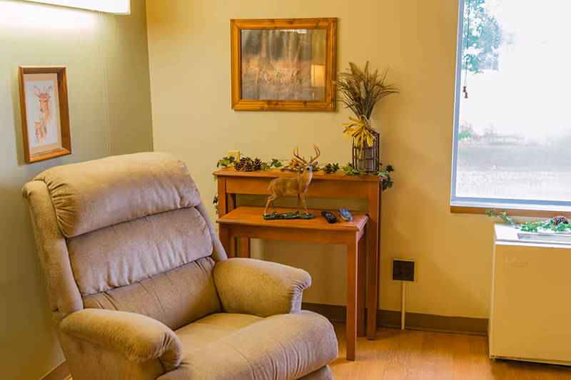 A cozy corner of a room featuring a beige recliner chair, two wooden nesting tables with a decorative deer figurine and remote controls, a framed deer artwork on the wall, a window letting in natural light, and a small white appliance near the window.