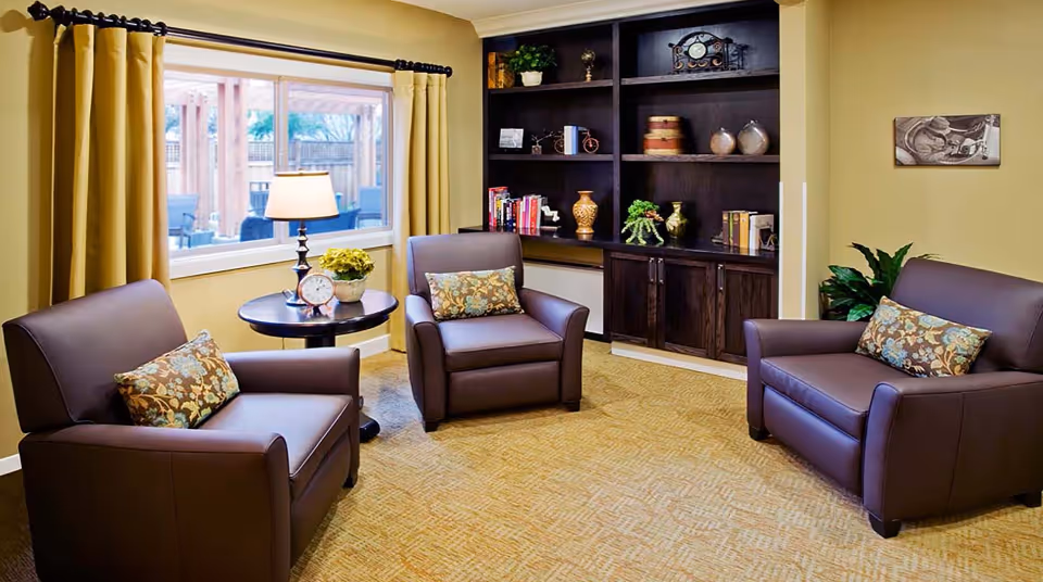 A cozy living room with three brown leather chairs, each with a decorative floral pillow. A round side table with a lamp, a clock, and a small plant sits near a window with yellow curtains. A dark wooden built-in bookshelf with various decorative items and books is against the wall. The room has beige walls and carpeted floor.
