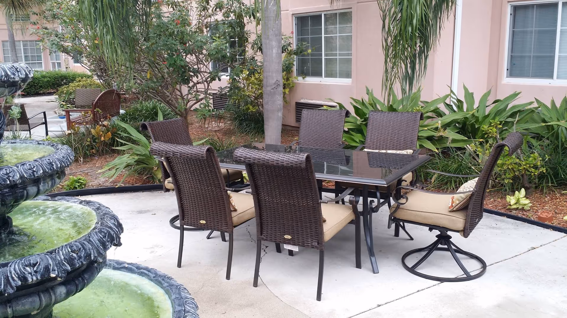 Outdoor patio area with a glass-top table surrounded by six cushioned wicker chairs. The patio is adjacent to a building with windows and surrounded by green plants and palm trees. A tiered water fountain is visible on the left side of the image.