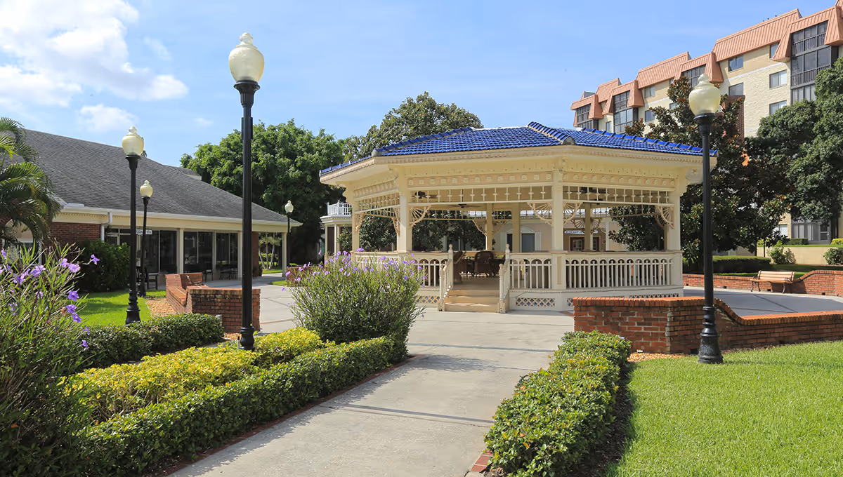 Outdoor view of a senior living facility featuring a white gazebo with a blue roof surrounded by green bushes and flowers. There are several black lamp posts with white globes lining the paved walkway leading to the gazebo. Buildings and trees are visible in the background under a partly cloudy sky.