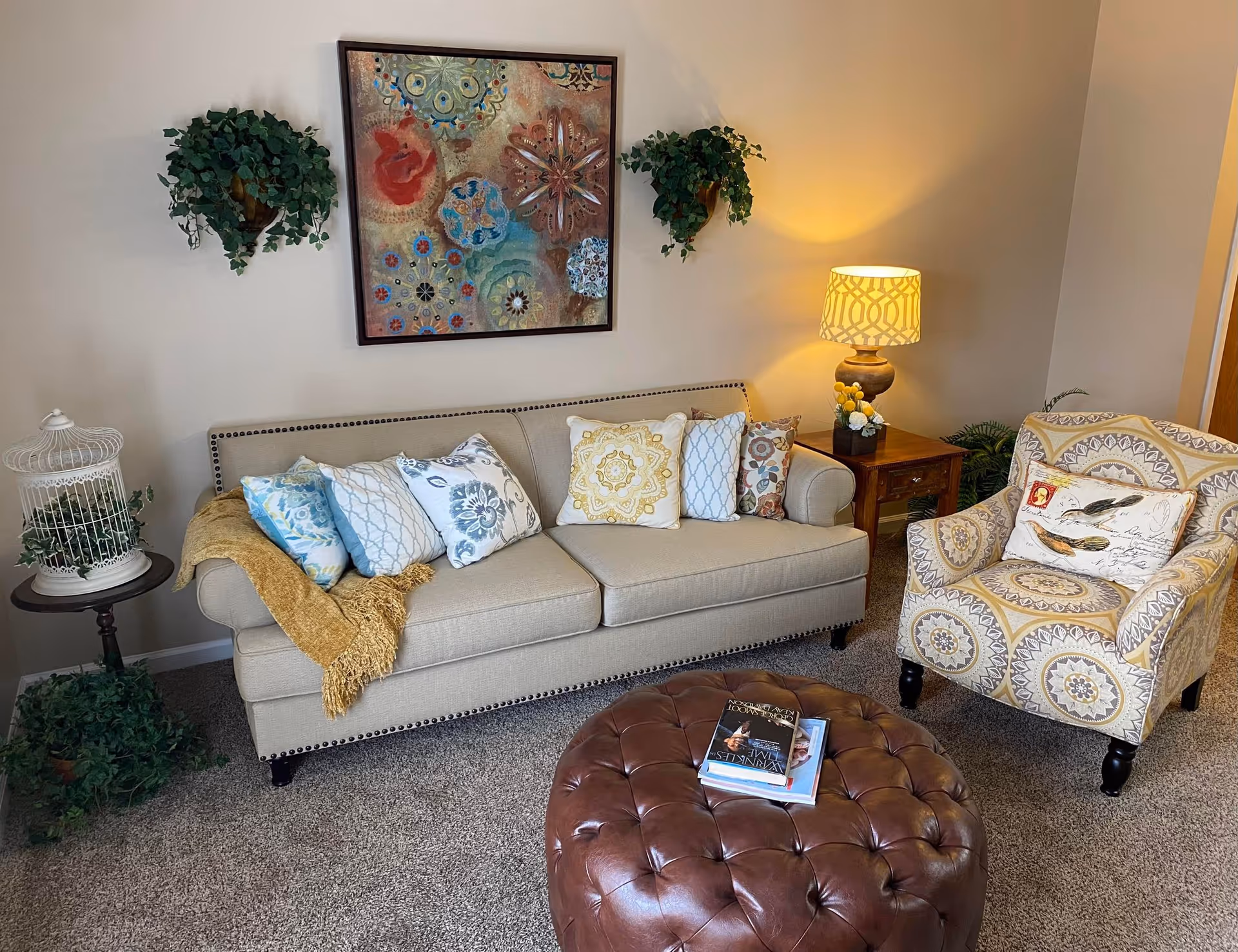 A cozy living room with a beige sofa adorned with multiple patterned pillows and a mustard yellow throw blanket. Next to the sofa is a small round table with a decorative birdcage and a potted plant. On the right side, there is a patterned armchair with a bird-themed pillow, a wooden side table with a lamp and a small floral arrangement. Two wall-mounted plants flank a colorful abstract painting above the sofa. A tufted brown leather ottoman with books on top is in the foreground on a carpeted floor.