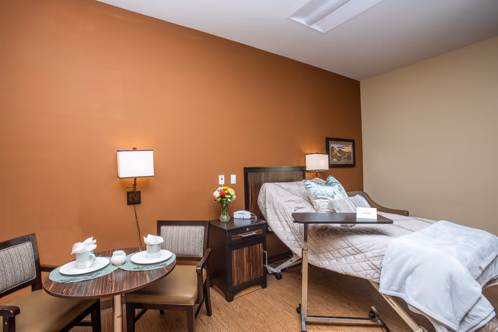 A neatly arranged hospital-style bedroom in a senior living facility, with a bed, two nightstands, a small round dining table with chairs, and an orange accent wall.