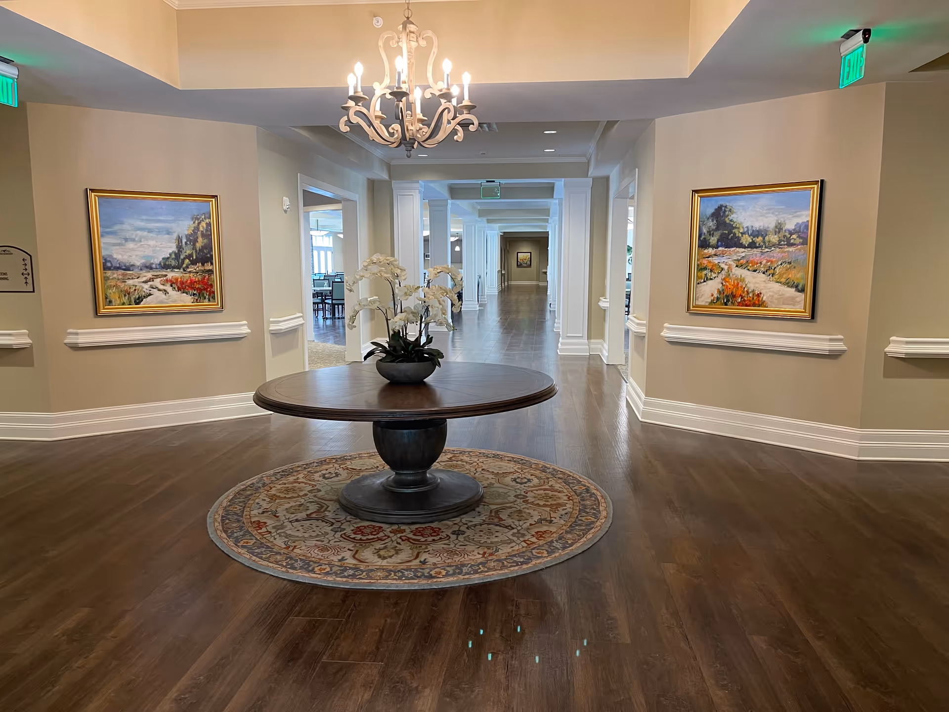 A spacious hallway in a senior living facility with wooden flooring, a round table with a flower arrangement on a circular rug in the center, two framed landscape paintings on the walls, and a chandelier hanging from the ceiling. The hallway leads to a dining area visible in the background.