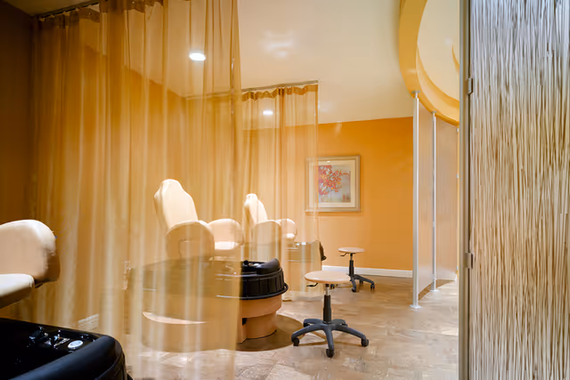 Interior view of a salon area with beige pedicure chairs, small rolling stools, and sheer golden curtains dividing the space. The walls are painted a warm orange color and a framed floral artwork is visible on the wall.