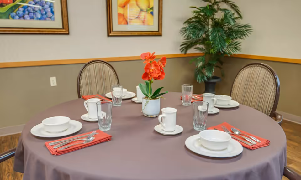 A round dining table set for six with white plates, bowls, cups, glasses, and silverware on red napkins. A small potted plant with red flowers is in the center. The room has two framed fruit paintings on the wall and a large potted plant in the corner.