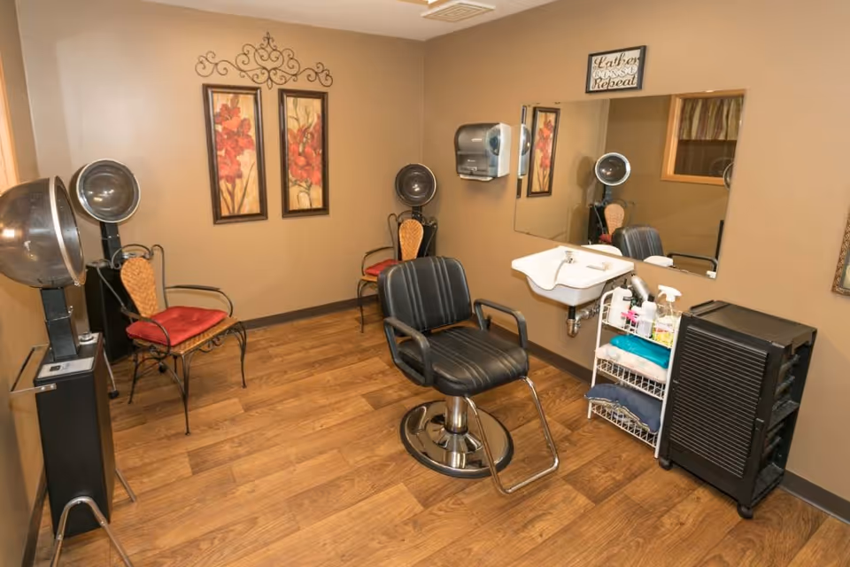 Interior view of a salon or hair styling room with a black salon chair in front of a wall-mounted sink and mirror. The room has wooden flooring, two vintage hair dryers, two decorative chairs with red cushions, and a small shelving unit with hair care products and towels. The walls are painted beige and decorated with two framed floral paintings and a decorative metal wall piece.