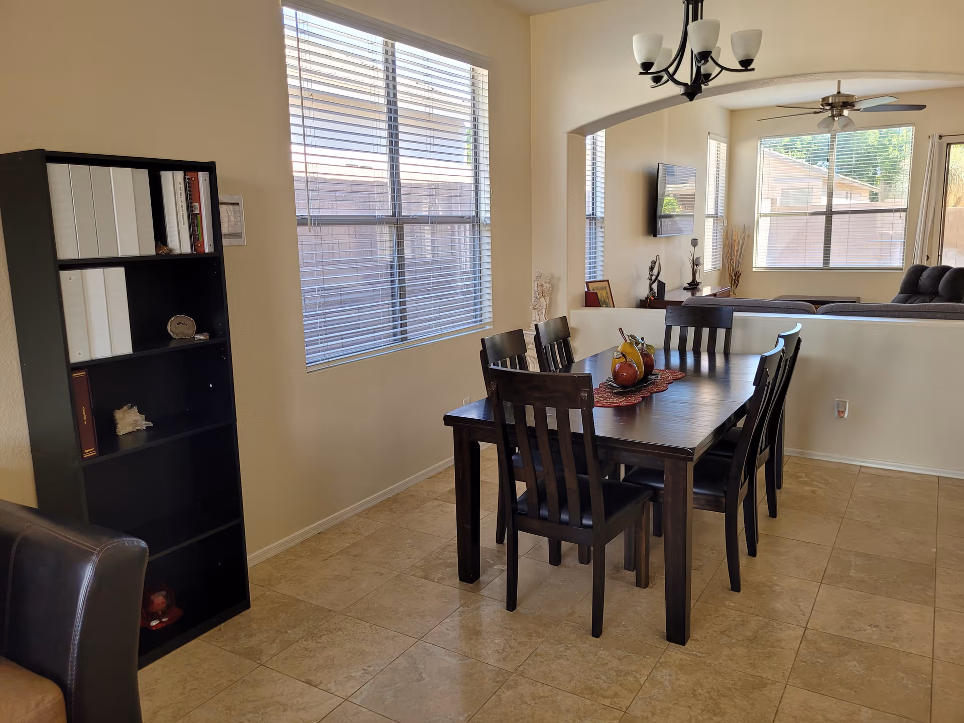Dining area with a dark wood table and six chairs, a fruit centerpiece, windows, a bookshelf, and a view into the living room.