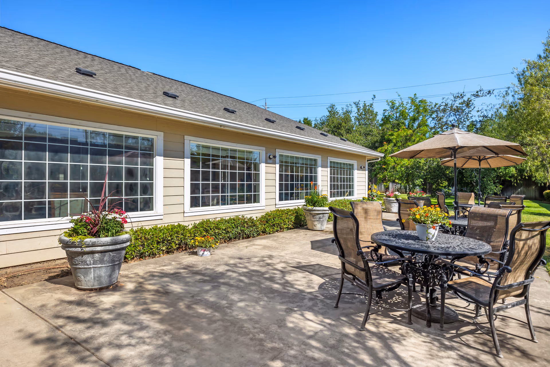 Outdoor patio area at Brookdale Lodi with metal tables and chairs, some shaded by large umbrellas, surrounded by potted plants and greenery under a clear blue sky.