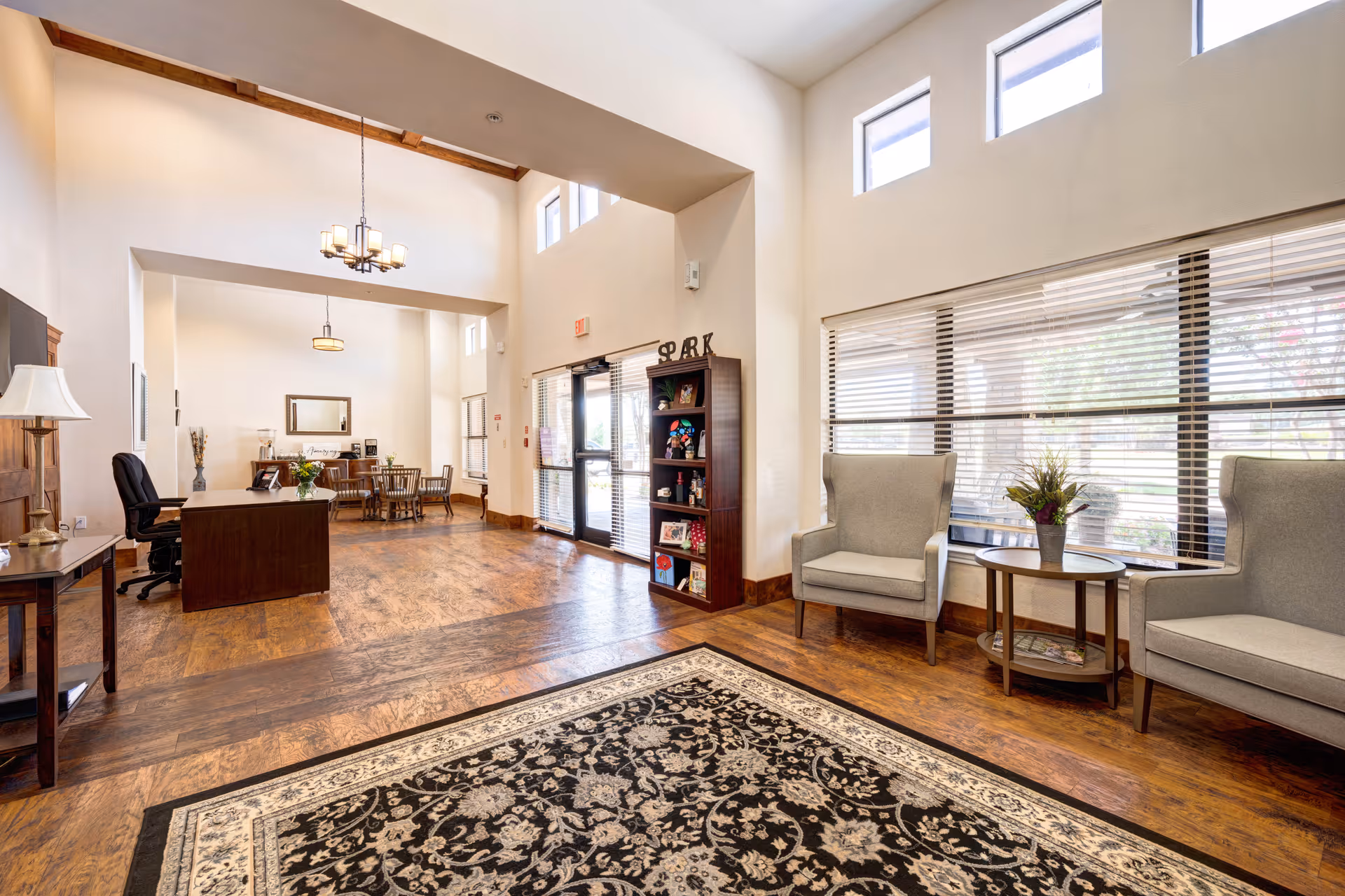A spacious and well-lit interior common area with wooden flooring and high ceilings. The room features a patterned area rug, two gray armchairs with a small round table between them, a wooden bookshelf with decorative items, a desk with an office chair, and a dining table with chairs in the background. Large windows with blinds allow natural light to fill the space.
