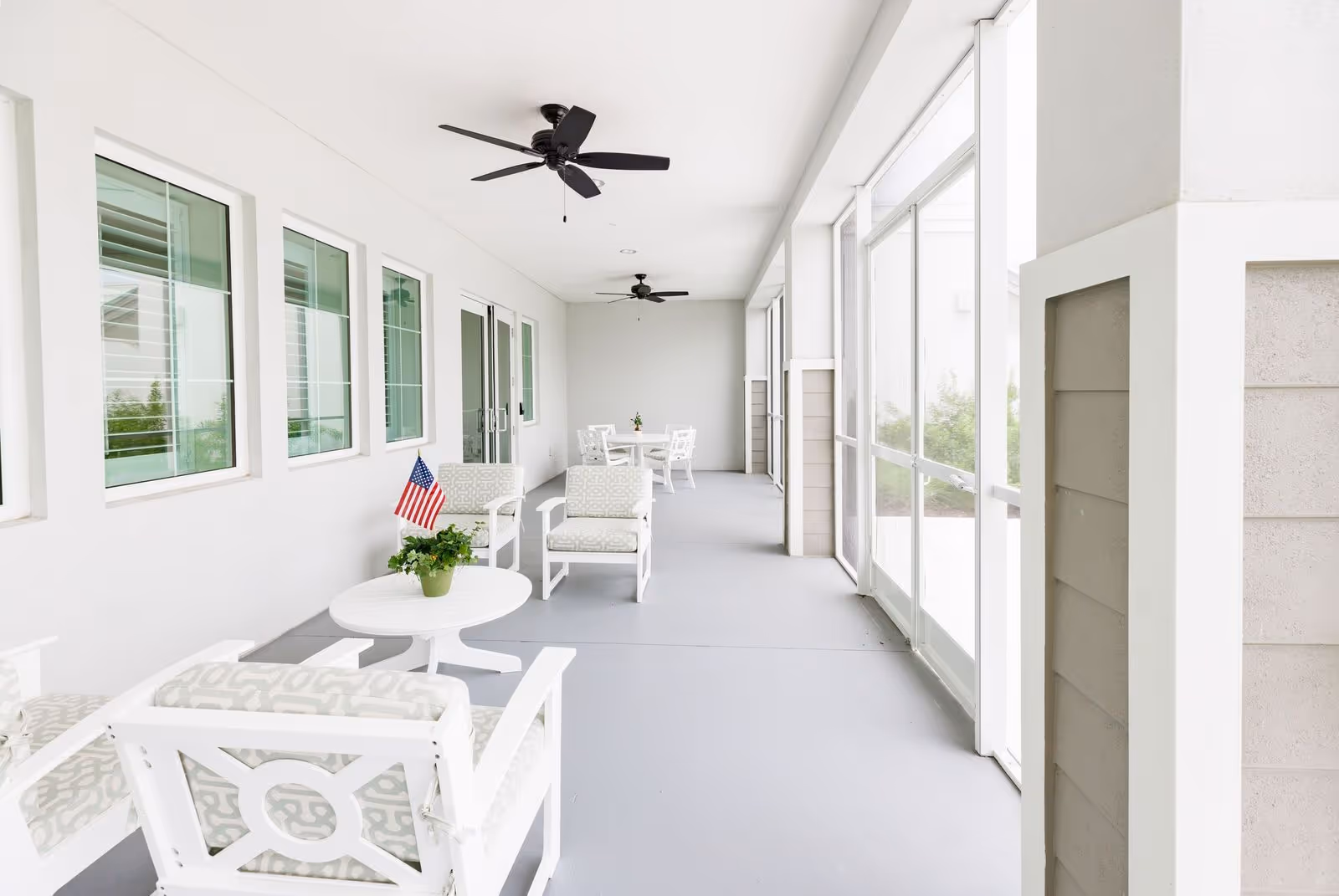 A bright, covered outdoor patio area with white cushioned chairs and small round tables. The patio has ceiling fans and large windows on one side, with a small American flag and a potted plant on one of the tables.