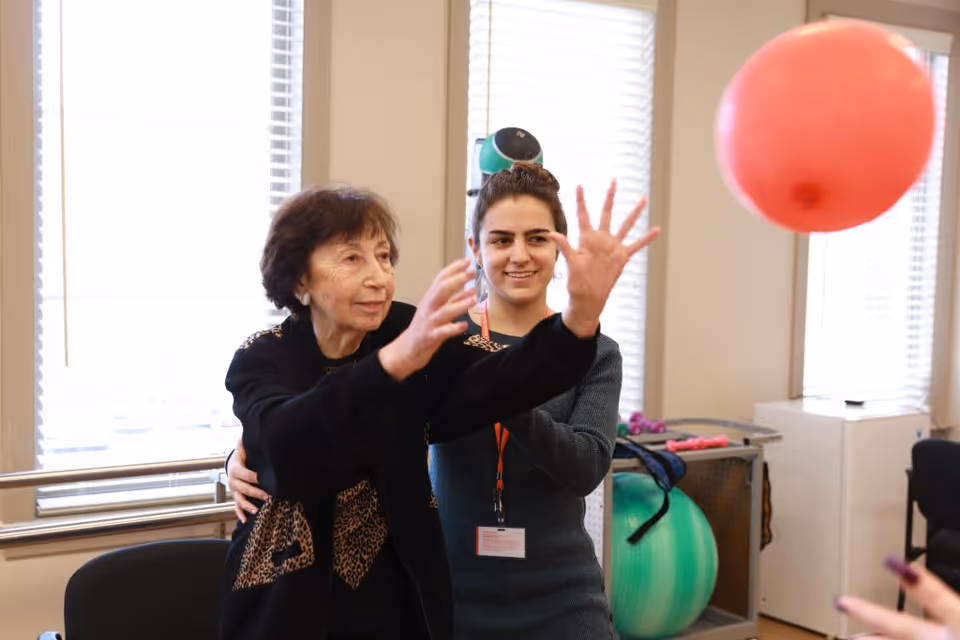An elderly woman is being assisted by a younger woman in a physical therapy or exercise session indoors. The elderly woman is reaching out to catch or throw a red balloon, while the younger woman supports her from behind. Exercise equipment like a green stability ball and small weights are visible in the background near windows with blinds.